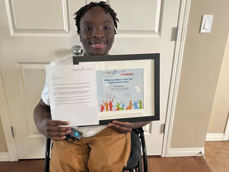 Saheed who is using his wheelchair holding his Difference Maker of the Year Award certificate. He is smiling and wearing tan-coloured pants.