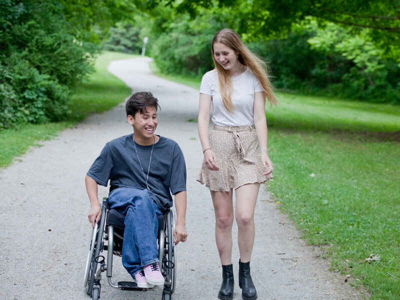 Two people talking and laughing while on a gravel path. One is using a wheelchair, has short brown hair and is wearing a dark t-shirt. The other person has long hair, is wearing a skirt and is walking.