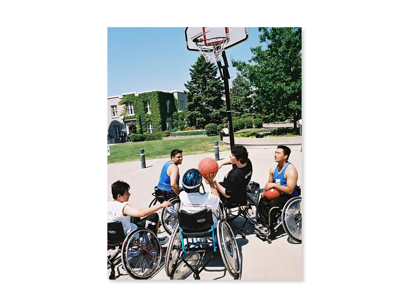 A group of five boys play wheelchair basketball outside.