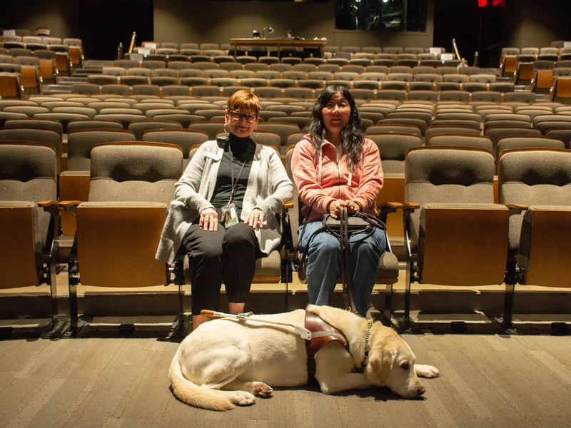 Two women sit in an empty auditorium, one of them accompanied by a guide dog lying calmly on the floor.