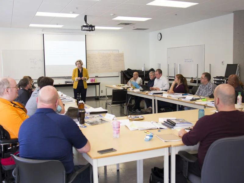 Group seated around tables in a classroom with attention to a presenter standing at the front near a projection screen and whiteboards. One participant is seated in a wheelchair, and notebooks, laptops, and water bottles are placed on the tables.