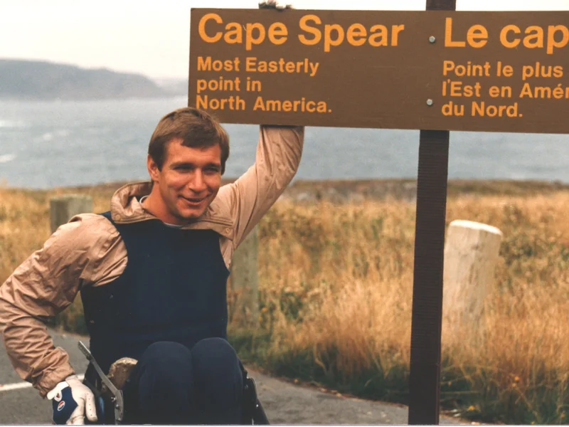 Rick Hansen au Cap Spear, à côté de l’enseigne « Le Cap Spear, l’endroit le plus à l’est du Canada ».