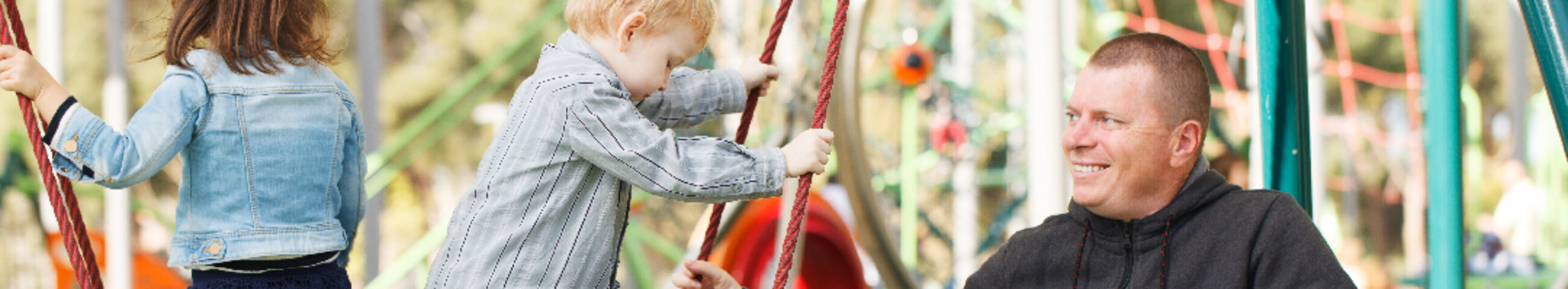 middle aged man and two young children in an accessible playground with adaptive equipment