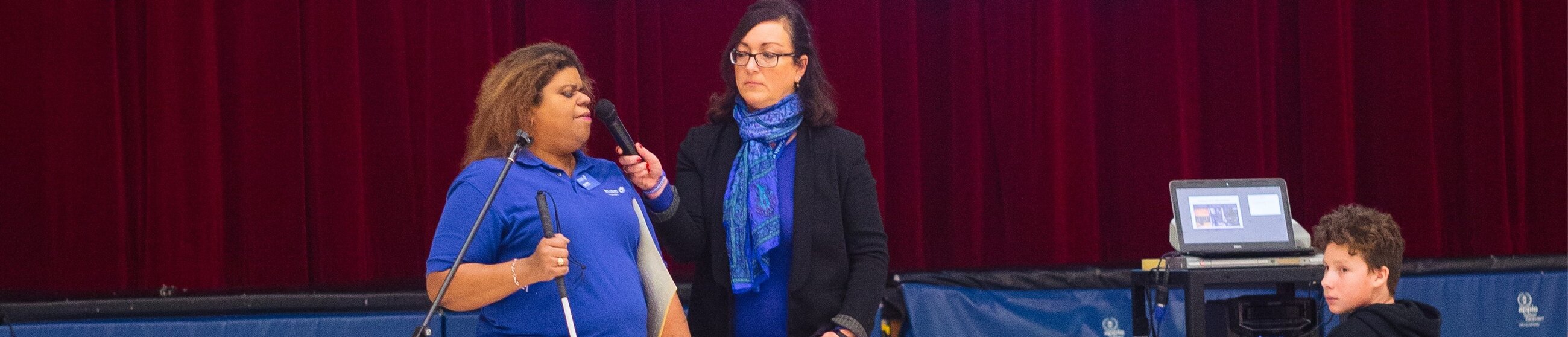 One person holds a white cane and a microphone, while the another person next to them speaks into a handheld microphone. A crowd of children is seated in the foreground.