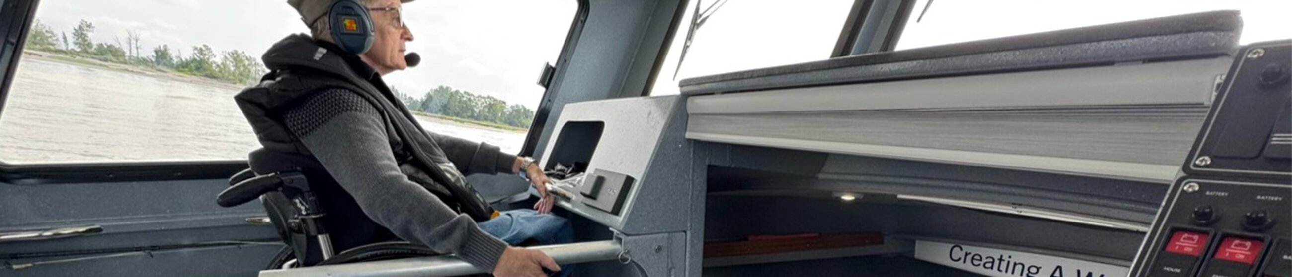 Person seated in a wheelchair inside a boat cabin, wearing a headset and holding the steering controls.