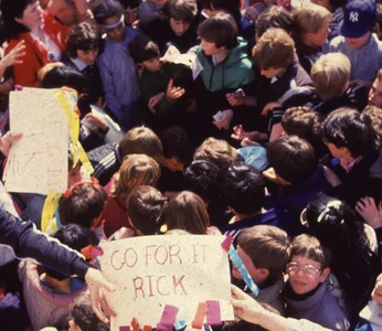 A crowd surrounds Rick Hansen, a sign says 'Go for it Rick' and one young teen looks up at the camera