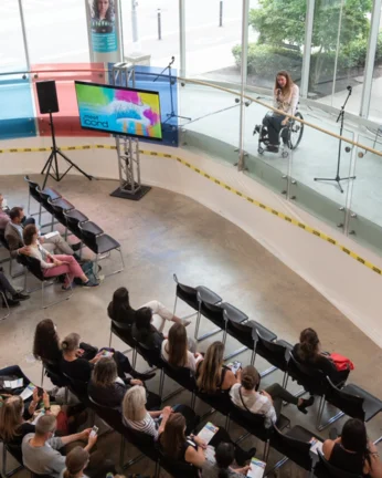 People gathered in an auditorium and on the podium a woman using a wheelchair