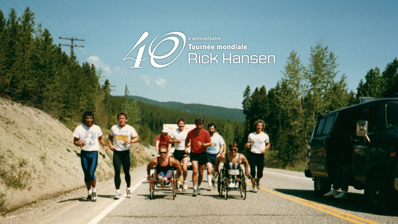Un groupe de personnes avance sur une route en milieu naturel, certaines en fauteuil roulant et d’autres en courant aux côtés. Le paysage est entouré de montagnes et d’arbres sous un ciel dégagé. Un texte superposé indique : « 40e anniversaire – Tournée mondiale Rick Hansen ».