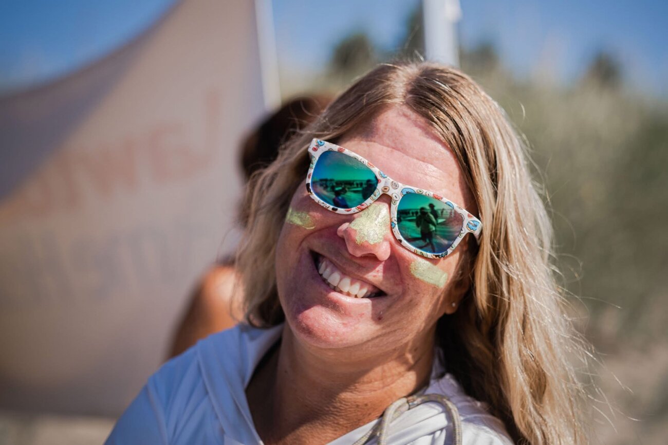 Close-up of a person outdoors wearing reflective sunglasses and face paint, smiling toward the camera. A large sculptural structure rises in the background against a clear blue sky.