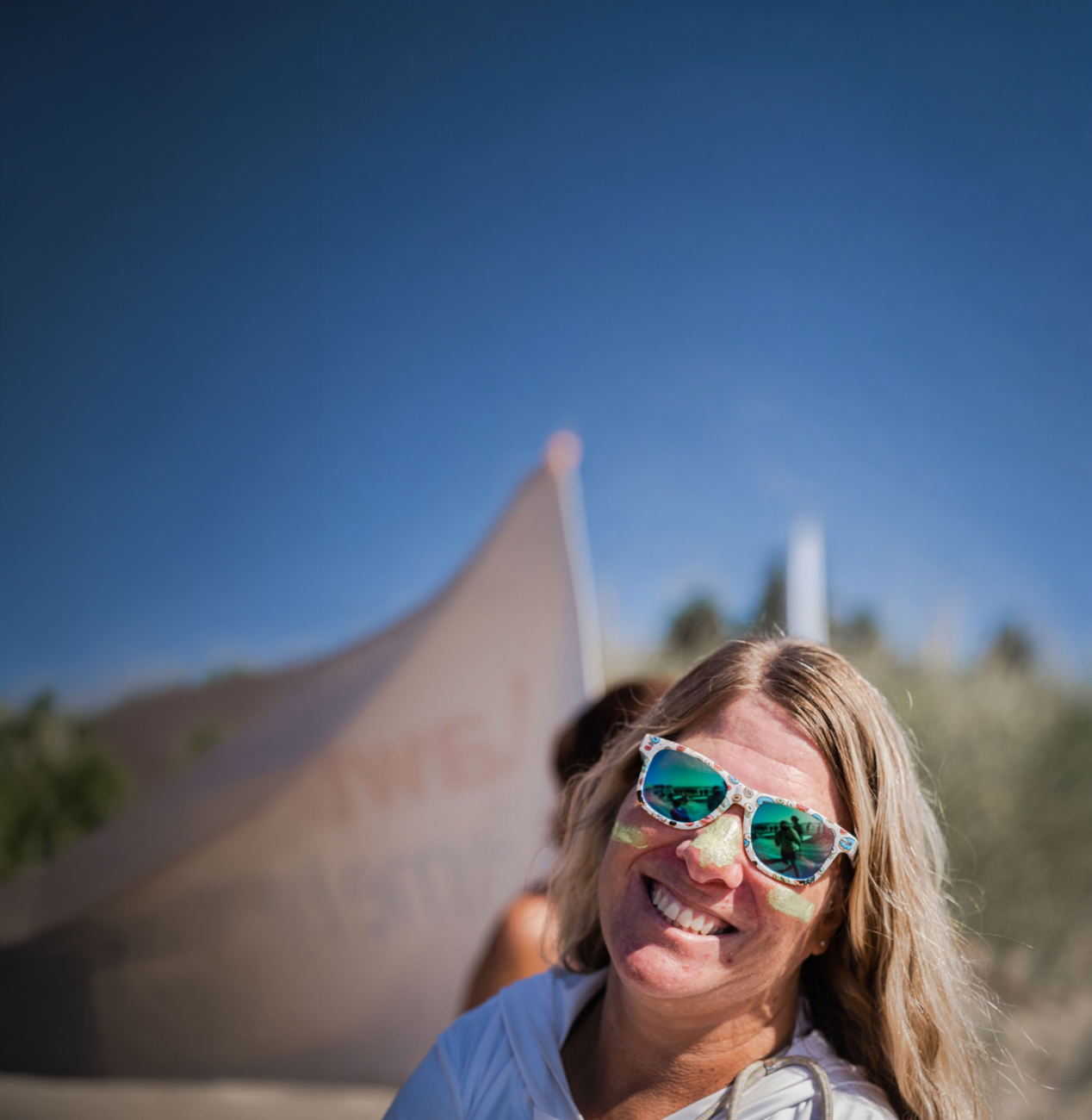 Close-up of a person outdoors wearing reflective sunglasses and face paint, smiling toward the camera. A large sculptural structure rises in the background against a clear blue sky.