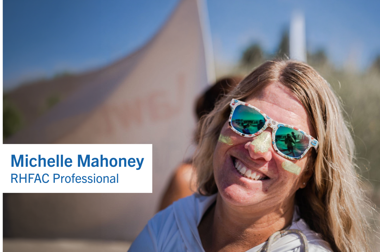 Woman outdoors wearing sunglasses and face paint, smiling toward the camera. A large sculptural structure rises in the background against a clear blue sky and a text that reads "Michelle Mahoney, RHFAC Professional."