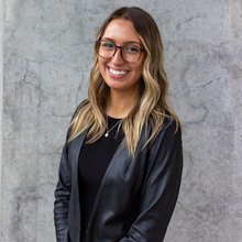 Woman with long blonde and brown hair with glasses smiling. She is wearing a black dress and a black leather blazer.