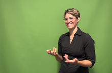 woman with short hair and glasses, smiling and signing ASL