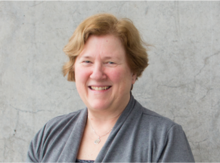 Middle aged women with short brown hair, blue eyes, smiling, wearing grey cardigan. 