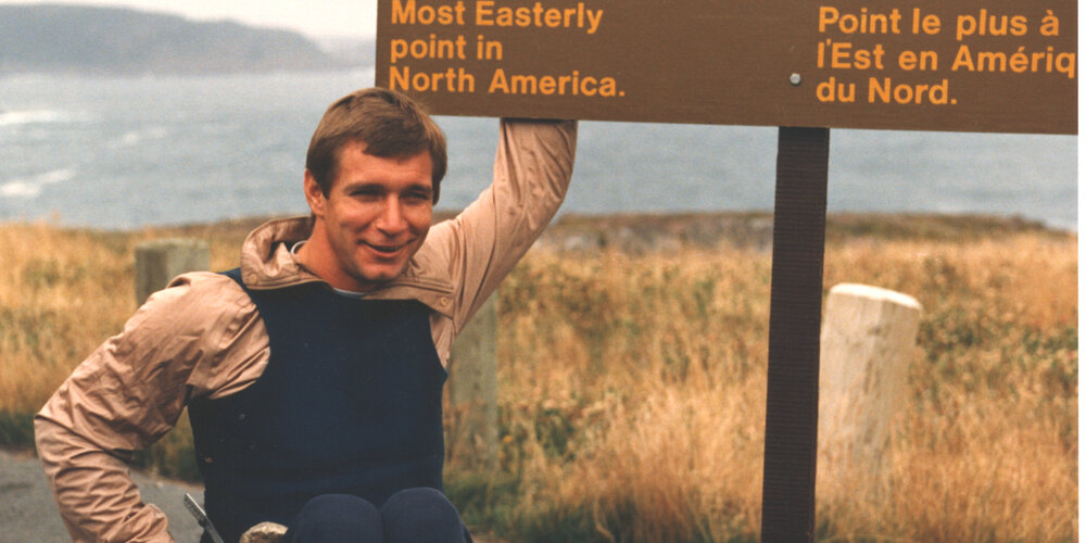 Rick Hansen au Cap Spear, à côté de l’enseigne « Le Cap Spear, l’endroit le plus à l’est du Canada ».