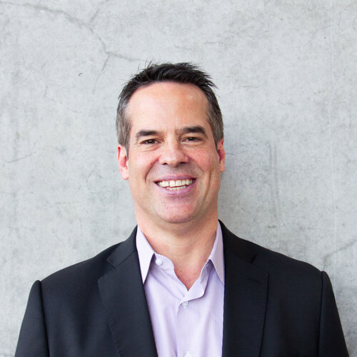 Middle aged man with short grey hair, brown eyes, smiling wearing a black blazer with lavender shirt.