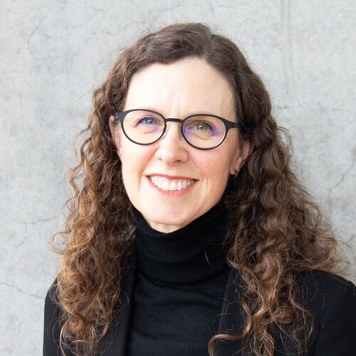 Woman with curly brown hair wearing glasses, smiling in a black turtleneck.