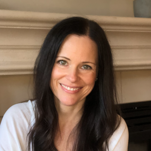 Woman with dark, long hair, smiling, wearing a white blouse, in front of a mantel.