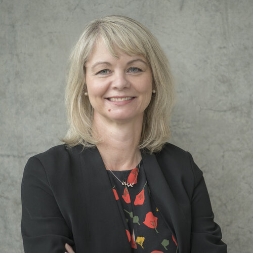 Woman with shoulder length blonde hair, smiling, dressed professionally with a blazer, against a stone background