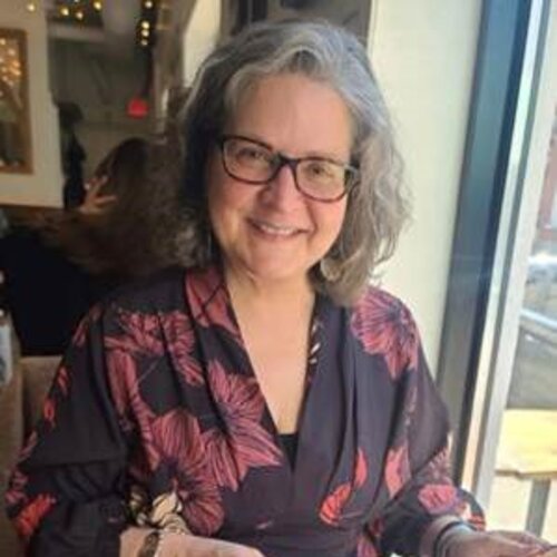grey haired woman with glasses wearing a floral blouse, smiling, sitting next to a window