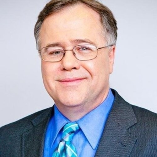 Man with short brown hair and glasses. He is wearing a blue dress shirt, a tie and a grey suit.