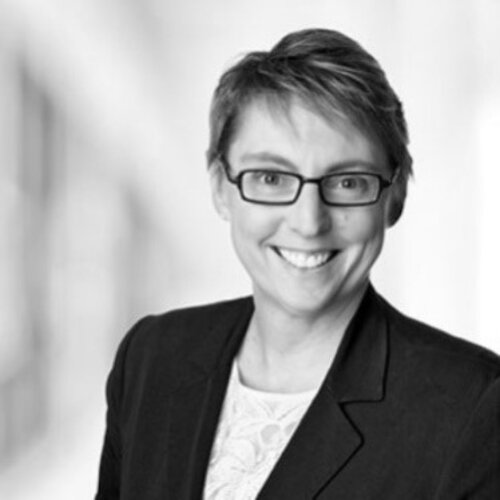 Black and white photo of a woman with short hair wearing glasses, a dark blazer and a white shirt. She is smiling.