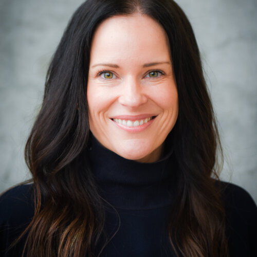 Woman with dark, long hair, smiling, wearing a black turtle neck sweater, in front of a grey background.