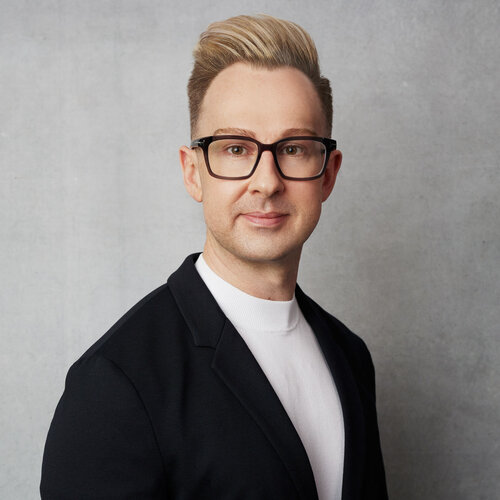 Jonathan Marriott who has short blonde hair, glasses and is wearing a dark suit jacket with a white shirt. He is in front of a grey background.