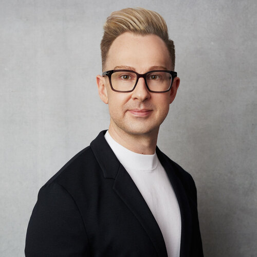 Jonathan Marriott who has short blonde hair, glasses and is wearing a dark suit jacket with a white shirt. He is in front of a grey background.
