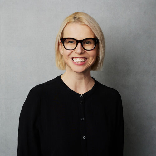 Sarah McCarthy who has chin-length blonde hair and is wearing thick black glasses and a black top. She is in front of a grey wall.