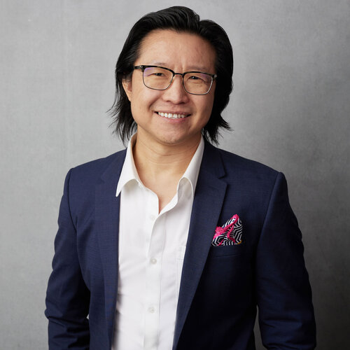 Kevin Ng, who has black hair and glasses and is standing in front of a grey wall. He is wearing a white button up shirt and a dark blue suit jacket.