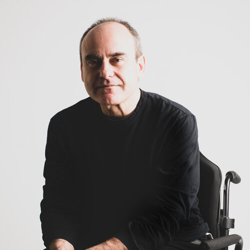 A headshot of John Ruffolo against a grey wall. He is wearing a black long-sleeve shirt and black pants, has short hair and is using a wheelchair.