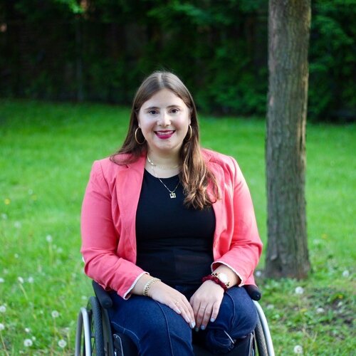 A woman with long brown hair, dressed in a pink blazer and black top, sits in a wheelchair in a grassy outdoor setting with trees in the background. She is smiling and looking at the camera.