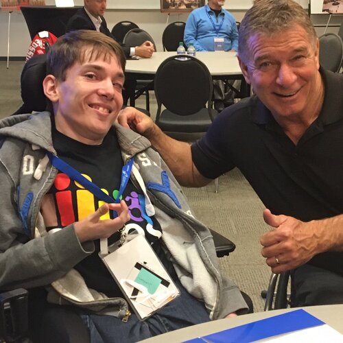Two smiling people in a classroom, both using wheelchairs
