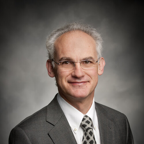 Middle aged man with grey short hair, wearing glasses, smiling, in a grey suit. 