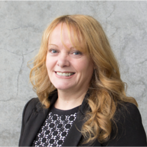  Middle aged woman with curly orange hair, blue eyes, smiling, wearing a black blazer with black patterned shirt. 