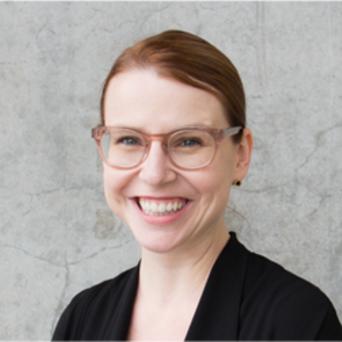 Woman with orange hair tied up, blue eyes, wearing glasses smiling in black blazer.