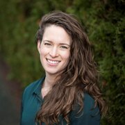 A woman with ling brown curly hair wearing a dark green shirt smiling.