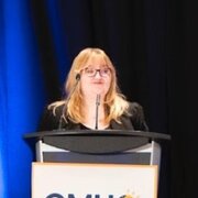 Young woman in a podium wearing glasses and speaking to the public
